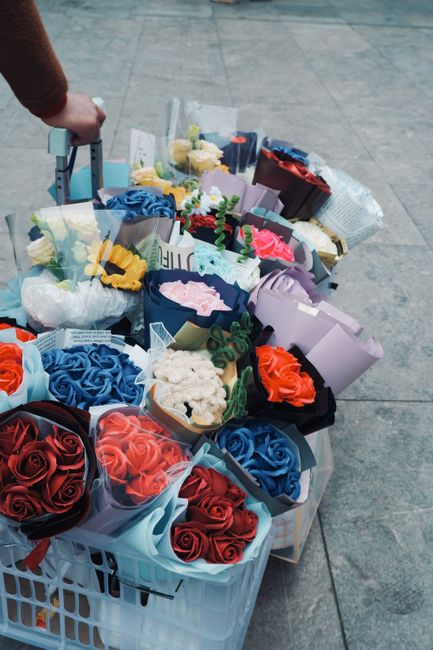 A collection of vibrant bouquet arrangements featuring red, blue, pink, white, and yellow roses displayed in various wrapping papers and decorative wraps, situated in a white plastic container on a pa