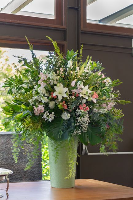 A tall, elegant floral arrangement placed in a green vase with a textured surface, featuring an assortment of fresh flowers including white lilies, light pink roses, and small white and pink filler bl