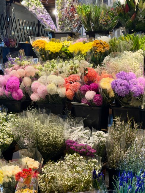 A vibrant display of fresh floral arrangements at a flower shop, featuring a variety of flowers in pastel pinks, purples, whites, and yellows. The bouquet arrangements include chrysanthemums, baby's b