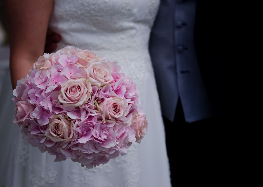 A close-up of a wedding bouquet held by a bride, consisting of soft pink roses and pink hydrangeas arranged in a rounded shape. The bouquet features fresh, delicate petals with visible texture and ste