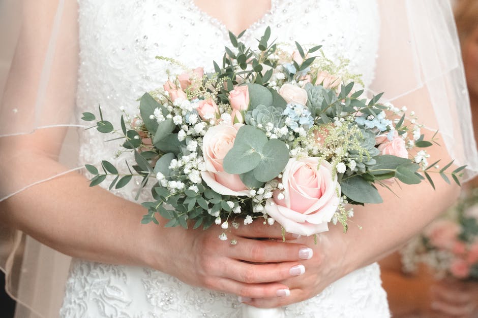 A bride in a white lace wedding dress is holding a round floral bouquet featuring soft pink roses, white baby’s breath, and sprigs of silver-green eucalyptus leaves. The bouquet is arranged in a com