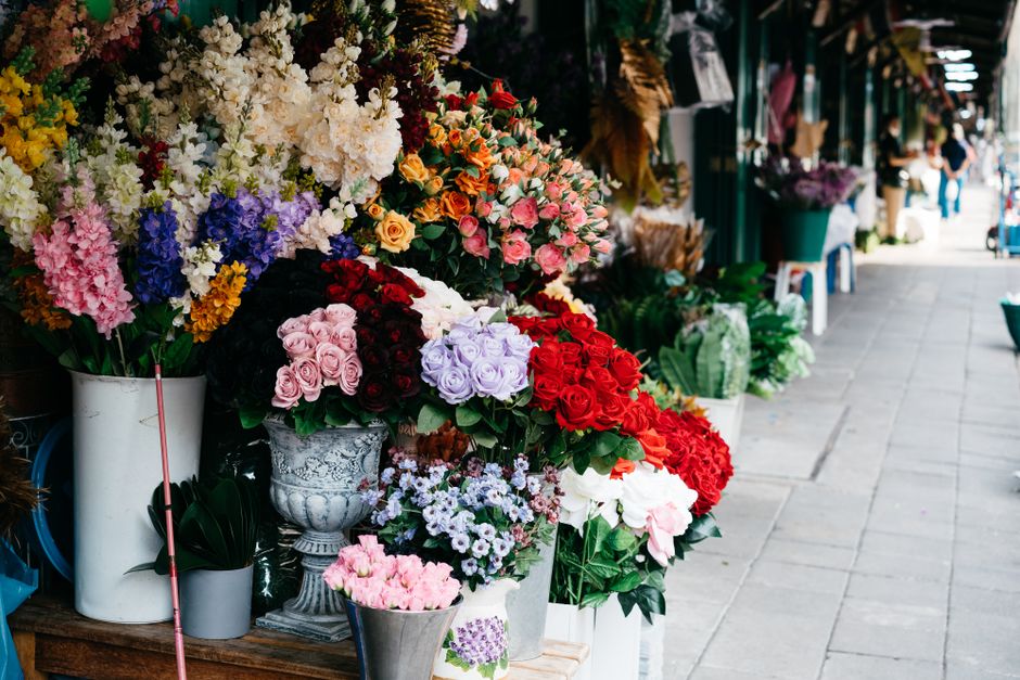 A display of various fresh flower bouquets arranged in white and decorative vases along a shopfront sidewalk. The selection includes roses, hydrangeas, and other blossoms in colors such as pink, red, 